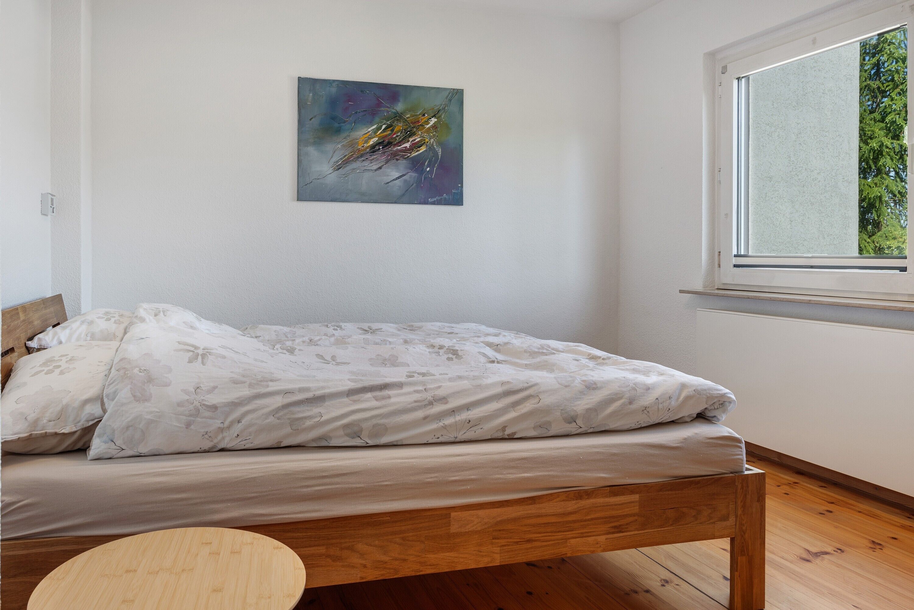 A bedroom with a bed, a round wooden table, a window, and a painting on the wall.
