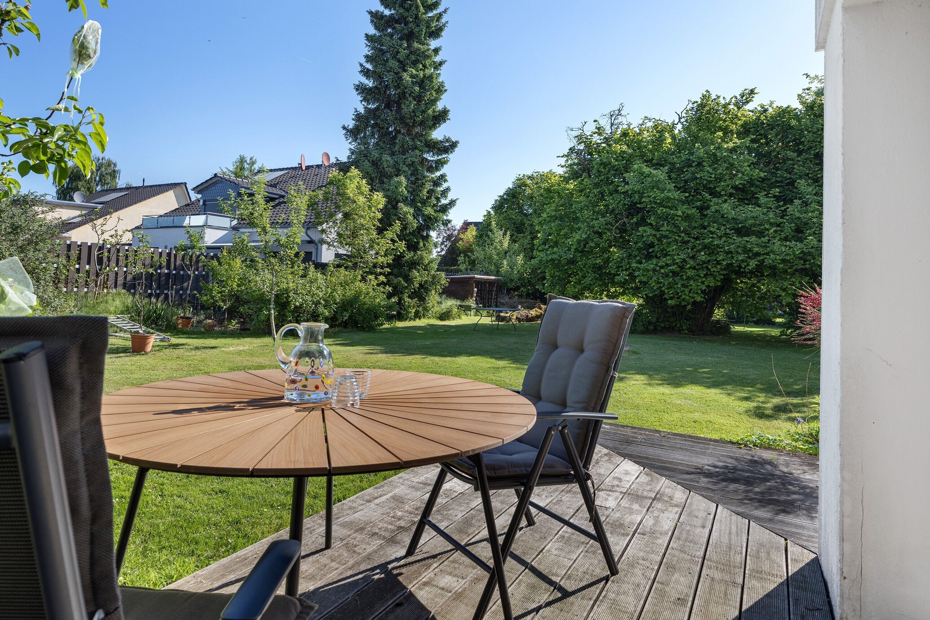 Eine Terrasse mit einem runden Holztisch und zwei Stühlen, einem Blumentopf und freiem Blick auf eine Wohngegend.