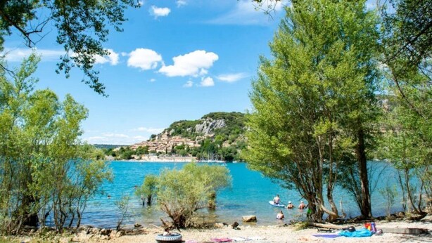 A beach with clear blue water, trees, and a small village in the background. - ZEcamping 3* chiens acceptés mobil-home clim 28m2 6 pers (Bauduen)