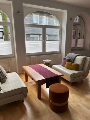 A modern living room with a sofa, a table, a round ottoman, and large windows with blinds.