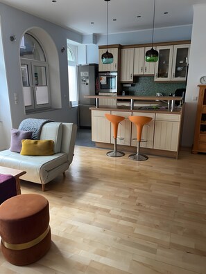 A modern kitchen with a central island, two bar stools, and a dining area with a sofa and ottoman.