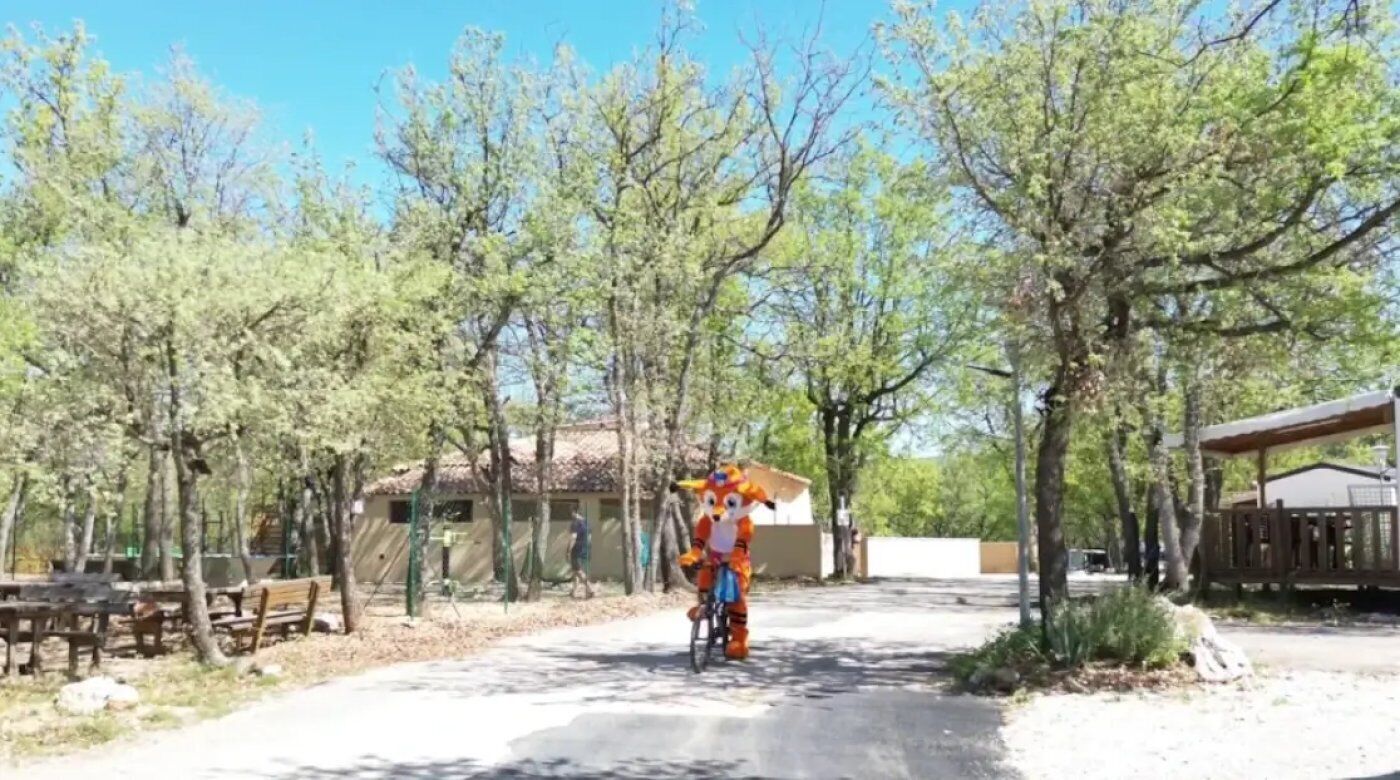 A paved pathway with a person on a bicycle, surrounded by trees and a building with a sloped roof.