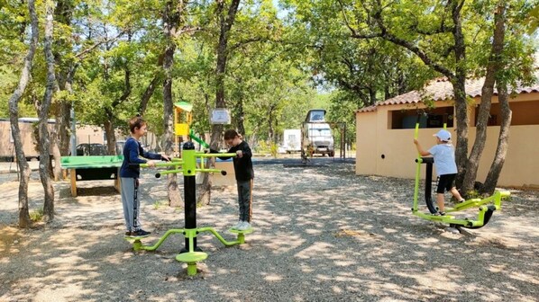 An outdoor exercise area with green fitness equipment, trees, and a building in the background.