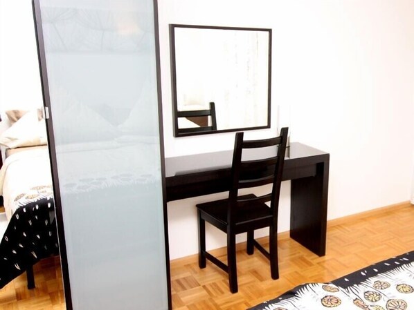 A room with a wooden floor, a black desk, a chair, a bed with patterned bedding, and a white wall.