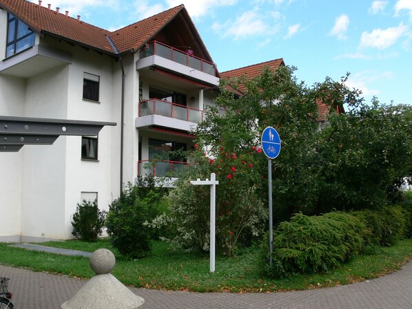 Un bâtiment moderne doté d’un balcon, d’une enseigne bleue indiquant un chemin piétonnier et une piste cyclable, et d’une zone pavée avec un poteau.