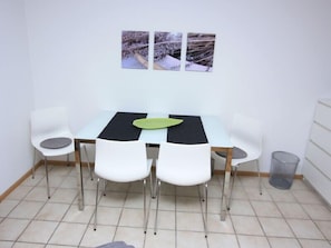 A dining area with a table, four chairs, a trash bin, and three framed pictures on the wall. - Apartment 'Ferienwohnung 2 Am Seepark Falk' with Balcony and Wi-Fi (Freiburg im Breisgau)