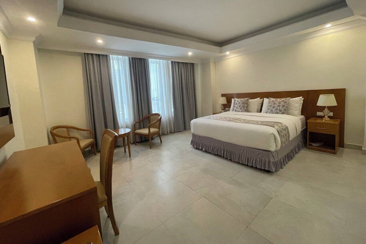 A hotel room with a large bed, a desk, two chairs, a nightstand with a lamp, and a window with curtains.