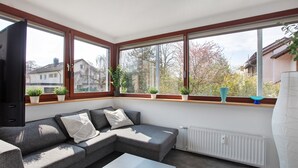 Un salon moderne avec un canapé d’angle gris, une table basse blanche et de grandes fenêtres ornées de plantes en pot.