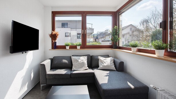 Un salon moderne avec un canapé d’angle, un téléviseur à écran plat fixé au mur et une grande fenêtre agrémentée de plantes en pot.