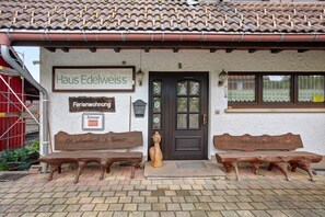Ein Gebäude mit einem Schild, auf dem 'Haus Edelweiss' steht, und einer Tür mit der Nummer 3. Vor dem Gebäude befinden sich zwei hölzerne Bänke.