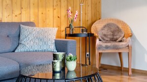 Una habitación con un sofá gris, una mesa redonda de vidrio con dos tazas metálicas y una pequeña planta en maceta, una silla de mimbre y una pared de madera.