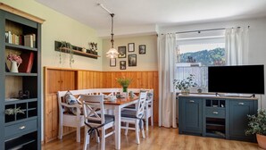 Un comedor con una mesa y sillas de madera, una estantería, un televisor sobre un mueble y una ventana con cortinas.