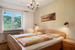A bedroom with a double bed, a nightstand, a bedside lamp, a window with curtains, a radiator, and a painting on the wall.