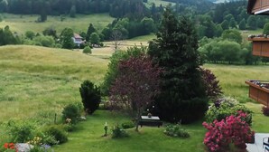 Paisaje rural con una casa, un jardín con diversas plantas y flores, y vistas de las colinas circundantes.