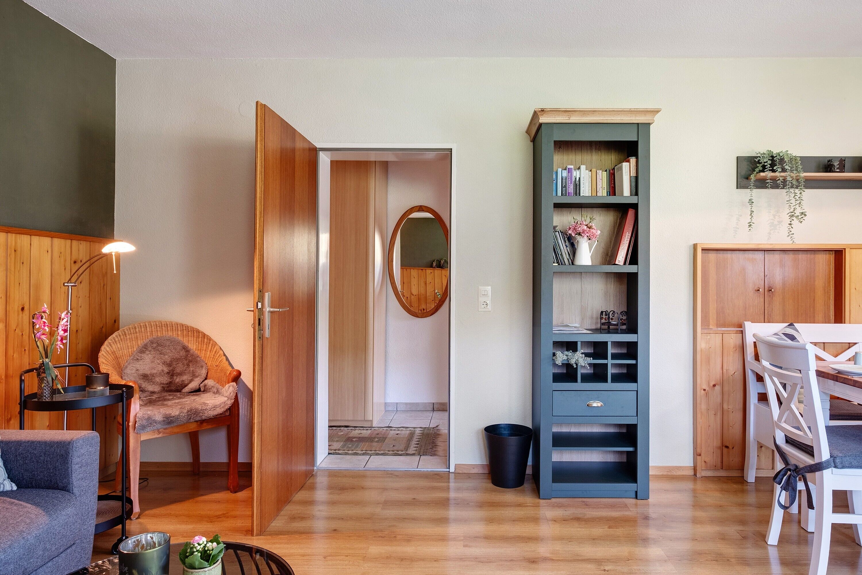 Una habitación con piso de madera, un sofá gris, una silla de mimbre, una estantería, una mesa de comedor con sillas y una puerta de madera que da a otra habitación.