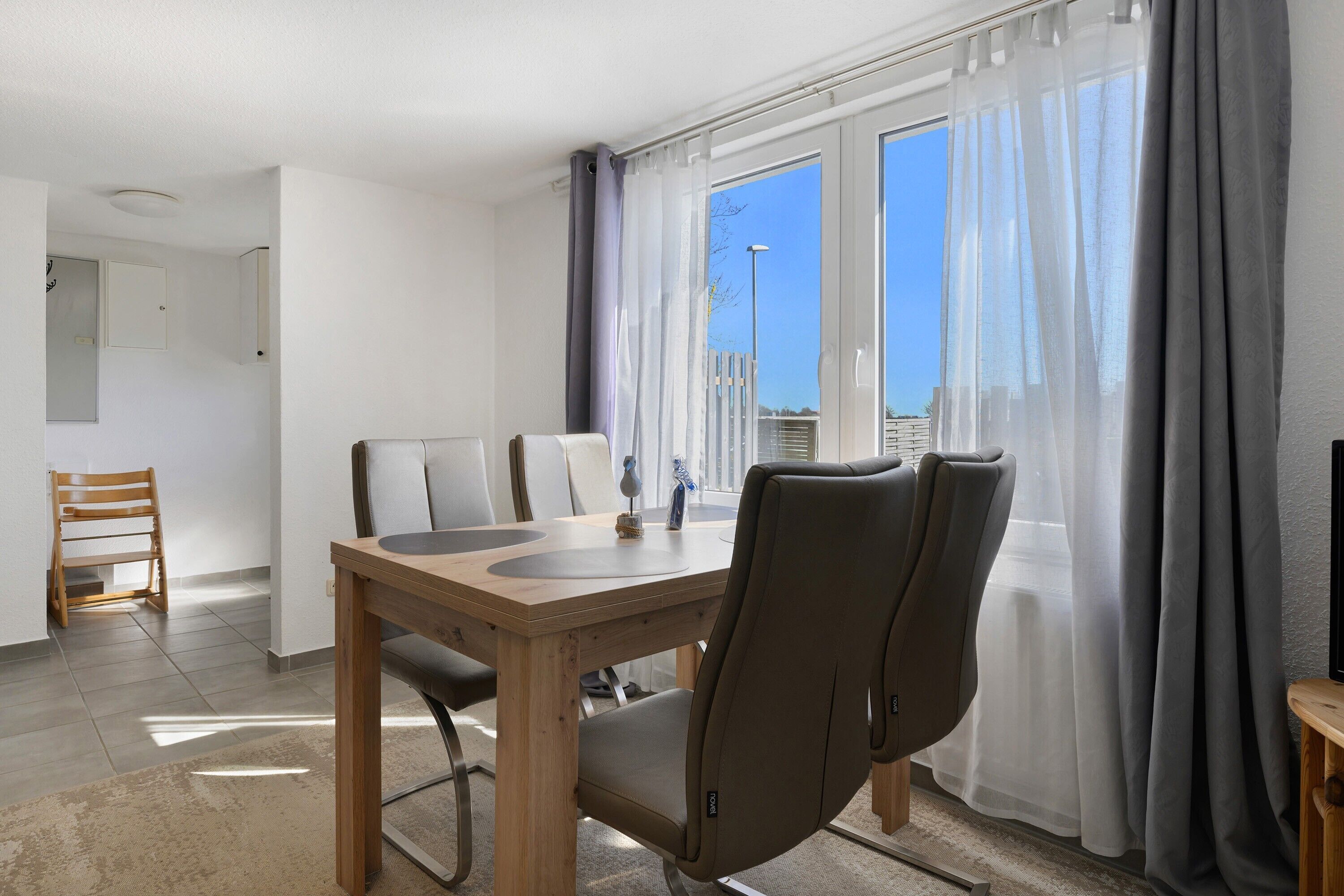 Una habitación con una mesa de madera, dos sillas, una mesita con un adorno y vistas amplias a través de ventanas con cortinas.