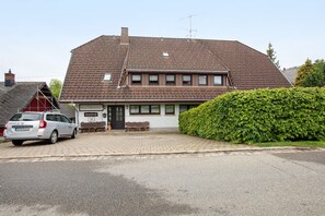 Une maison Ă deux Ă©tages, avec un toit en tuiles, un banc devant lâentrĂ©e et une voiture garĂ©e dans lâallĂ©e.