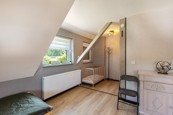 A room with a sloped ceiling, a crib, a chair, a radiator, and a window with a view of greenery.