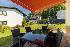 Un patio con una mesa de vidrio y sillas de mimbre, un columpio y una casa al fondo.
