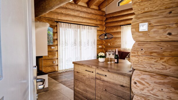 A log cabin interior with wooden walls, a wooden counter with a vase and wine glasses, and a window with curtains.