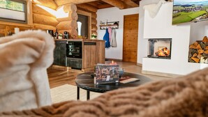 A cozy living room with a fireplace, wooden walls, a kitchen area, and a view of the countryside.