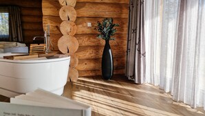 A wooden cabin interior with a white bathtub, a wooden wall with circular cutouts, a vase with greenery, and curtains.