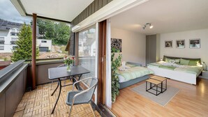 A balcony with a table and chairs, a bed with a green and white bedspread, and a view of the outside. - Apartment Schanzenblick with Sauna & Pool in the Black Forest (Schönwald im Schwarzwald)
