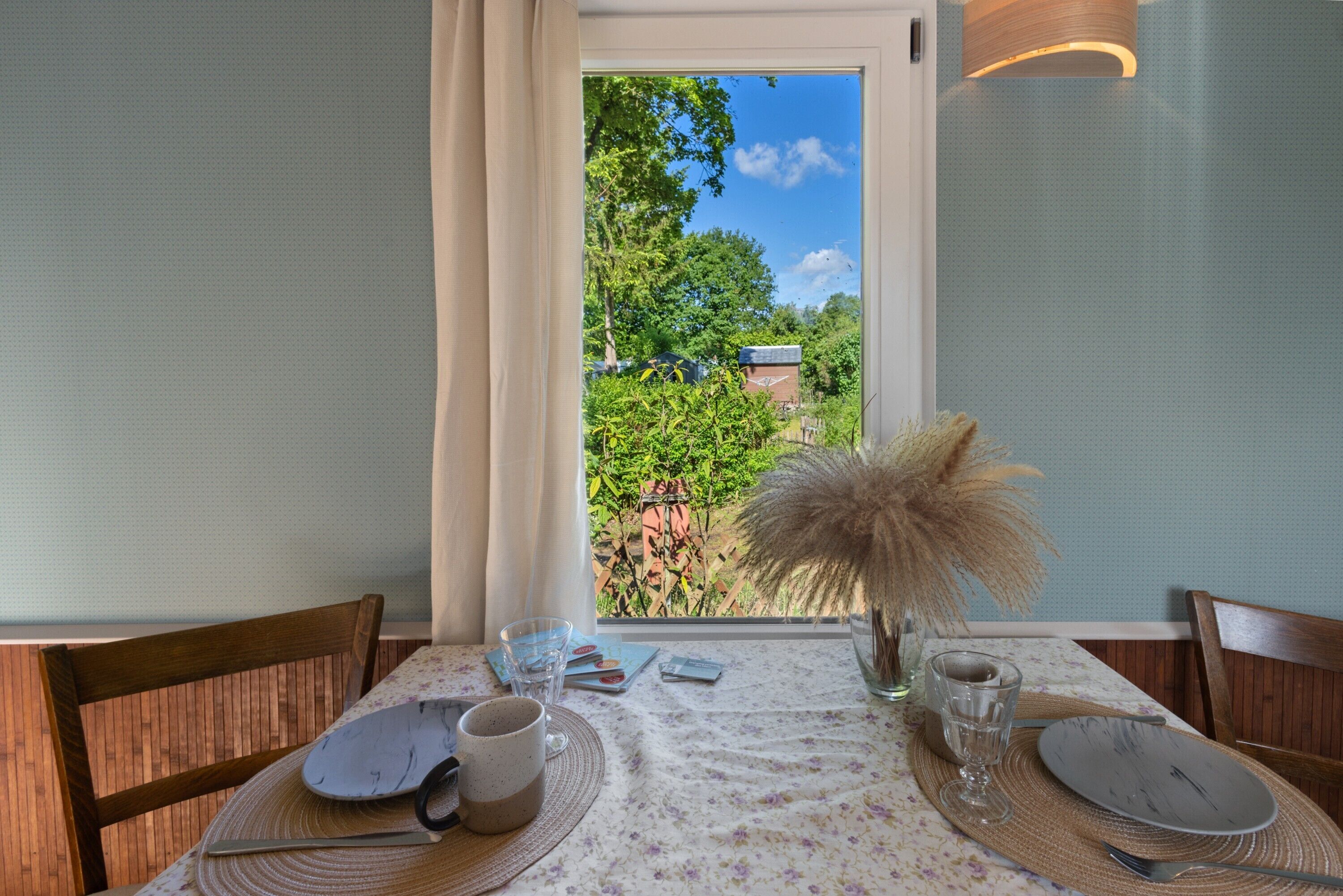 A dining table with a floral tablecloth, a vase with dried pampas grass, and a view of greenery outside.