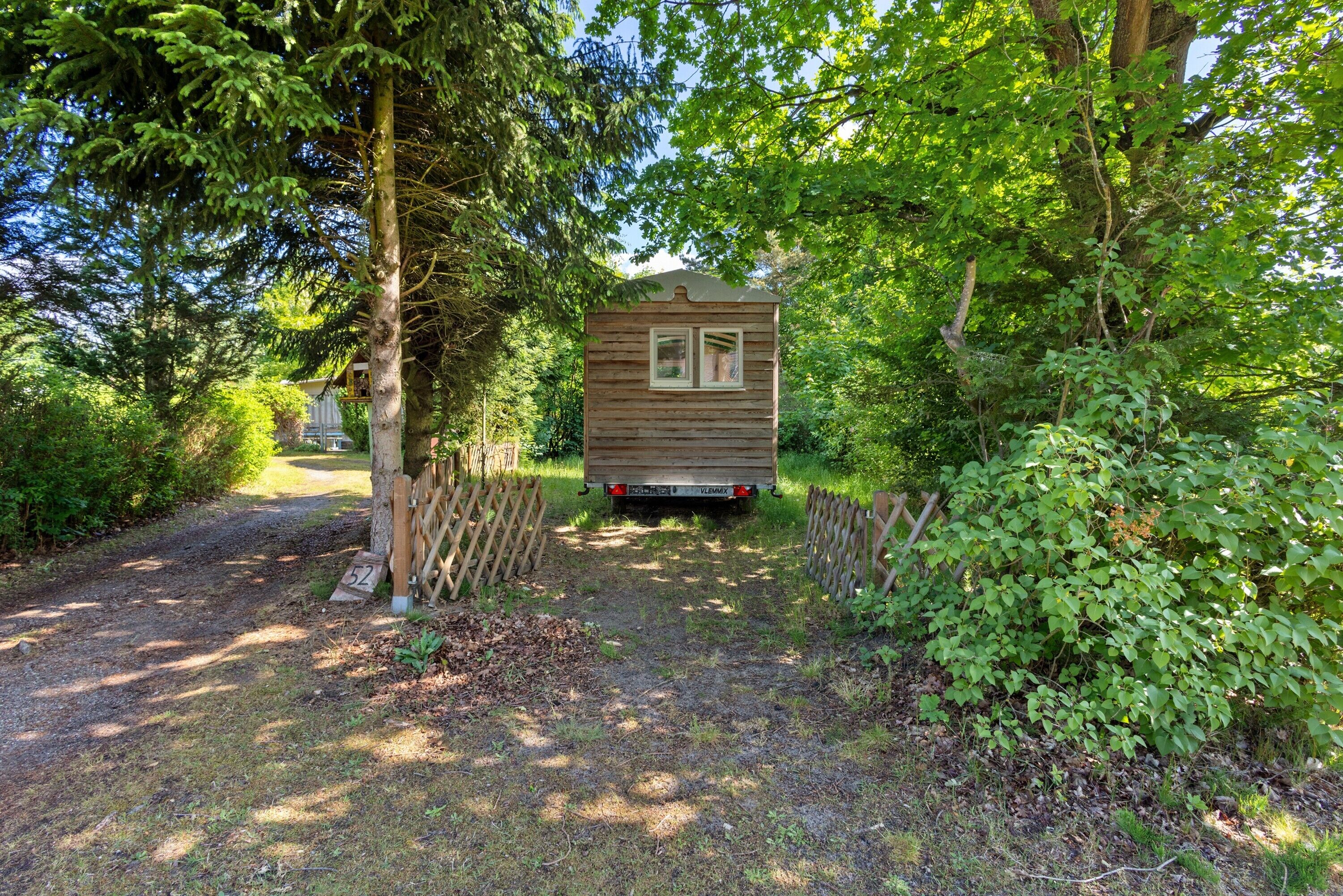 A small wooden cabin with a window, surrounded by dense greenery and a gravel path leading to it.