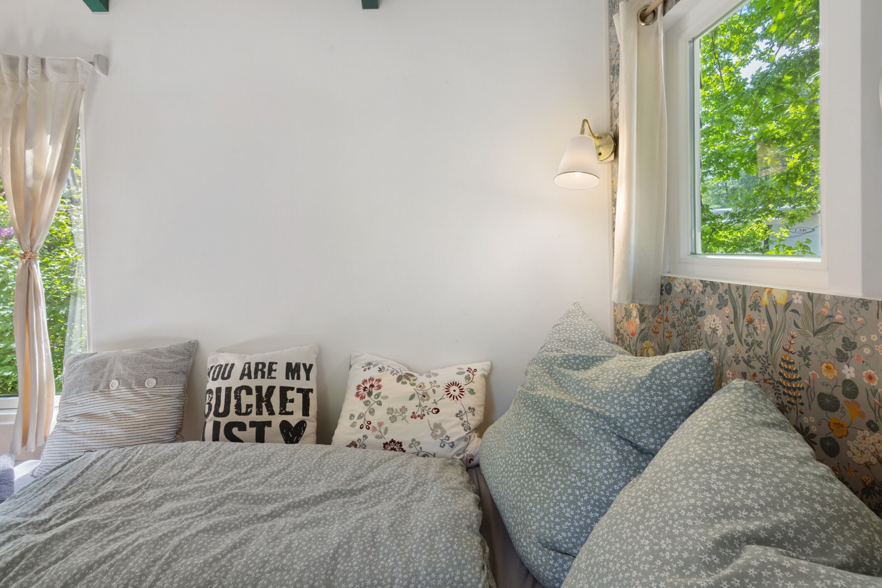 A bedroom with a bed, pillows, a window with a view, and a wall-mounted light.