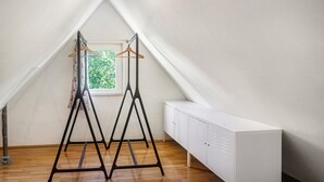 Ein Zimmer mit schrägem Dach, einem Fenster mit Blick auf Grünflächen, einem weißen Schrank und einem schwarzen Metallgestell mit Kleiderbügeln.