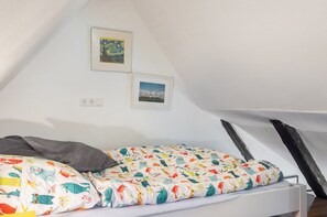 Un pequeño dormitorio abuhardillado con forma de triángulo, una cama, dos cuadros enmarcados y techo blanco.