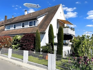 Une maison à deux étages avec un toit en pente, un balcon orné de plantes en pot et une clôture blanche.