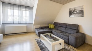 Un salon moderne avec un plafond en pente, un canapé d’angle gris, une table basse en verre et un tableau représentant un paysage urbain accroché au mur.