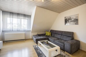 Un salon moderne avec un plafond en pente, un canapé d’angle gris, une table basse en verre et un tableau représentant un paysage urbain accroché au mur.