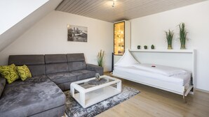 Una sala moderna con un sofá gris, una mesa de centro blanca y una cama también blanca. Se aprecia un cuadro de paisaje urbano en la pared y una planta sobre la cama.