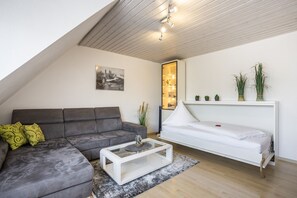 Een moderne woonkamer met een grijze bank, een witte salontafel en een wit bed. Aan de muur hangt een stadsgezicht schilderij en er staat een plant op het bed.