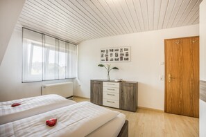 Une chambre avec deux lits, une armoire en bois, un mur décoré d’un collage de photos encadrées et une fenêtre ornée de rideaux légers.