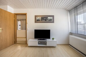 Un salon moderne avec un téléviseur à écran plat posé sur un meuble blanc, une photographie encadrée représentant un paysage urbain accrochée au mur et une porte en bois donnant sur une autre pièce.