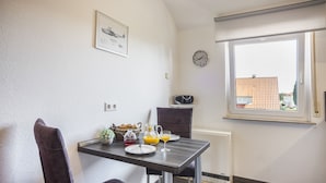 Un coin repas avec une table dressée pour deux personnes, une horloge au mur et une fenêtre donnant sur des immeubles.
