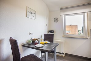 Un coin repas avec une table dressée pour deux personnes, une horloge au mur et une fenêtre donnant sur des immeubles.