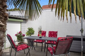 Een terras buiten met een tafel en stoelen, omringd door potplanten en een witte muur.