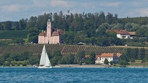 A coastal scene with a sailboat, a large building with a tower, and vineyards. - Apartment 'Bodensee' with Garden, Balcony and Wi-Fi (Friedrichshafen)