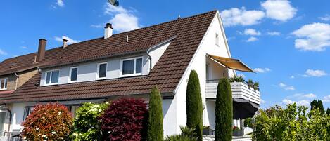 A two-story house with a brown roof, white walls, and a balcony with potted plants.