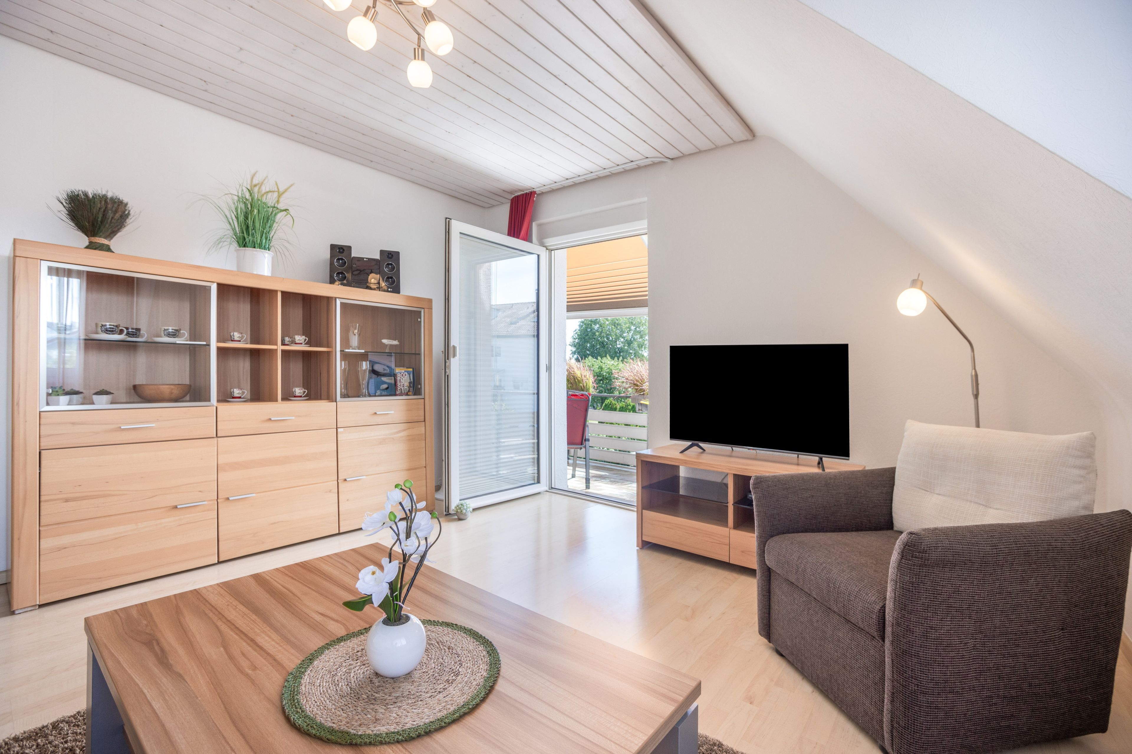 Ein modernes Wohnzimmer mit einem Holzkabinett, einem Couchtisch mit Blumenvase, einem Fernseher auf einem Standfuß und einer Couch.