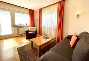 Ein Wohnzimmer mit Sofa, Sessel und Couchtisch, einem Fenster mit Vorhängen sowie einer Wandleuchte.