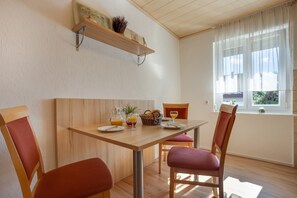 Een eetgedeelte met een houten tafel, rode fauteuils, een raam met dunne gordijnen en een plank met een vaas en een bordje.