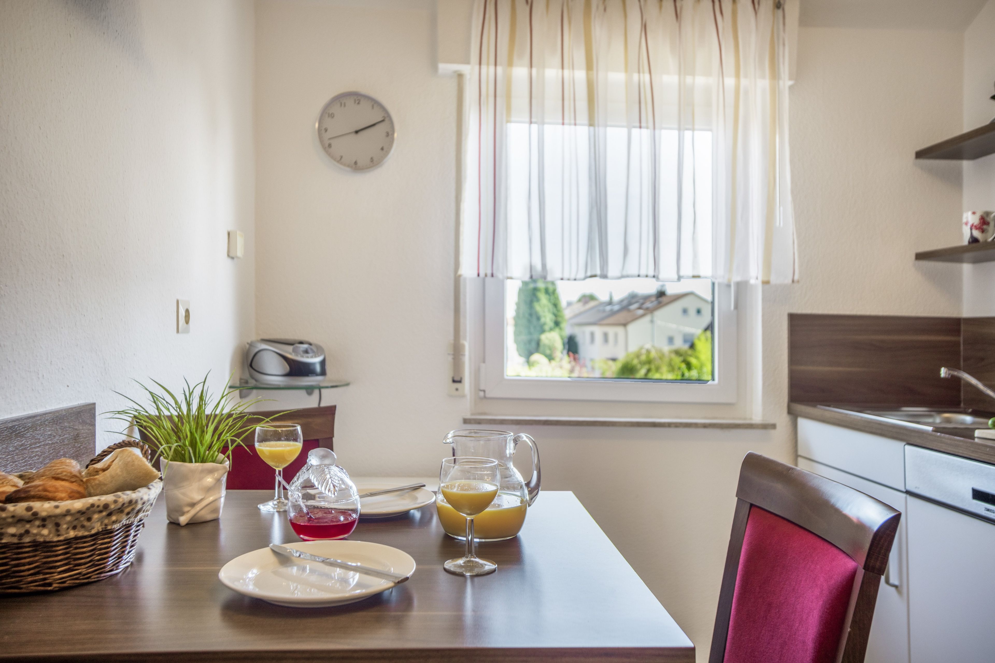 A dining area with a table set for two, a clock on the wall, and a window with a view of houses.