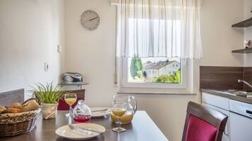 A dining area with a table set for two, a clock on the wall, and a window with a view of houses.