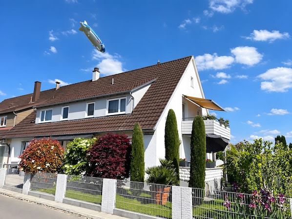 A two-story house with a brown roof, white walls, and a balcony with potted plants.